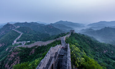 Great Wall segments winding across green mountains under a hazy blue sky with distant ridges.