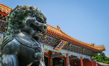 Forbidden City, Beijing, Chinatravel, Bronze guardian lion statue in front of a traditional Chinese temple with ornate, colorful roof tiles against a clear blue sky.