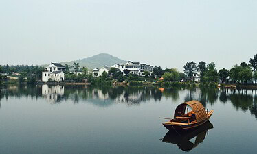 Chinatrip, Calm lakeside scene with white houses along the shore, green trees, and a wooden boat in the foreground on a reflective lake.