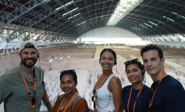 Terra Cotta Warriors, xi'an, china, china tour, Five people smiling for a selfie at an indoor archaeological dig, with a large curved roof and excavated area behind them, all wearing orange lanyards and headsets.