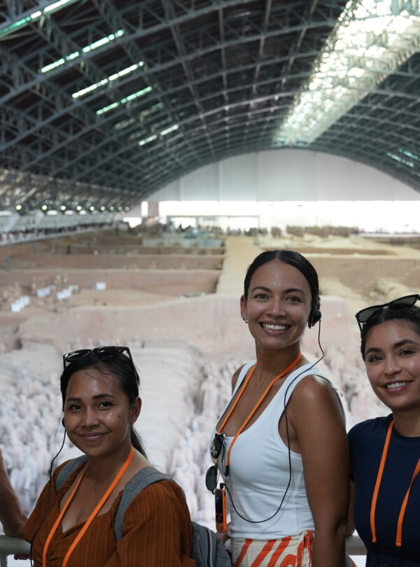 Terra Cotta Warriors, xi'an, china, china tour, Five people smiling for a selfie at an indoor archaeological dig, with a large curved roof and excavated area behind them, all wearing orange lanyards and headsets.