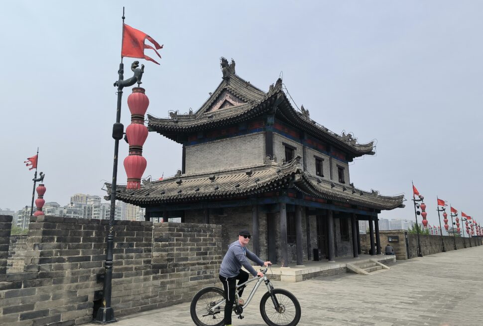 Tourists cycling atop Xi'an's historic City Wall, Chinatravel, Traditional Chinese temple-like building with ornate curved roof, red lanterns, and a cyclist passing by along a stone wall.