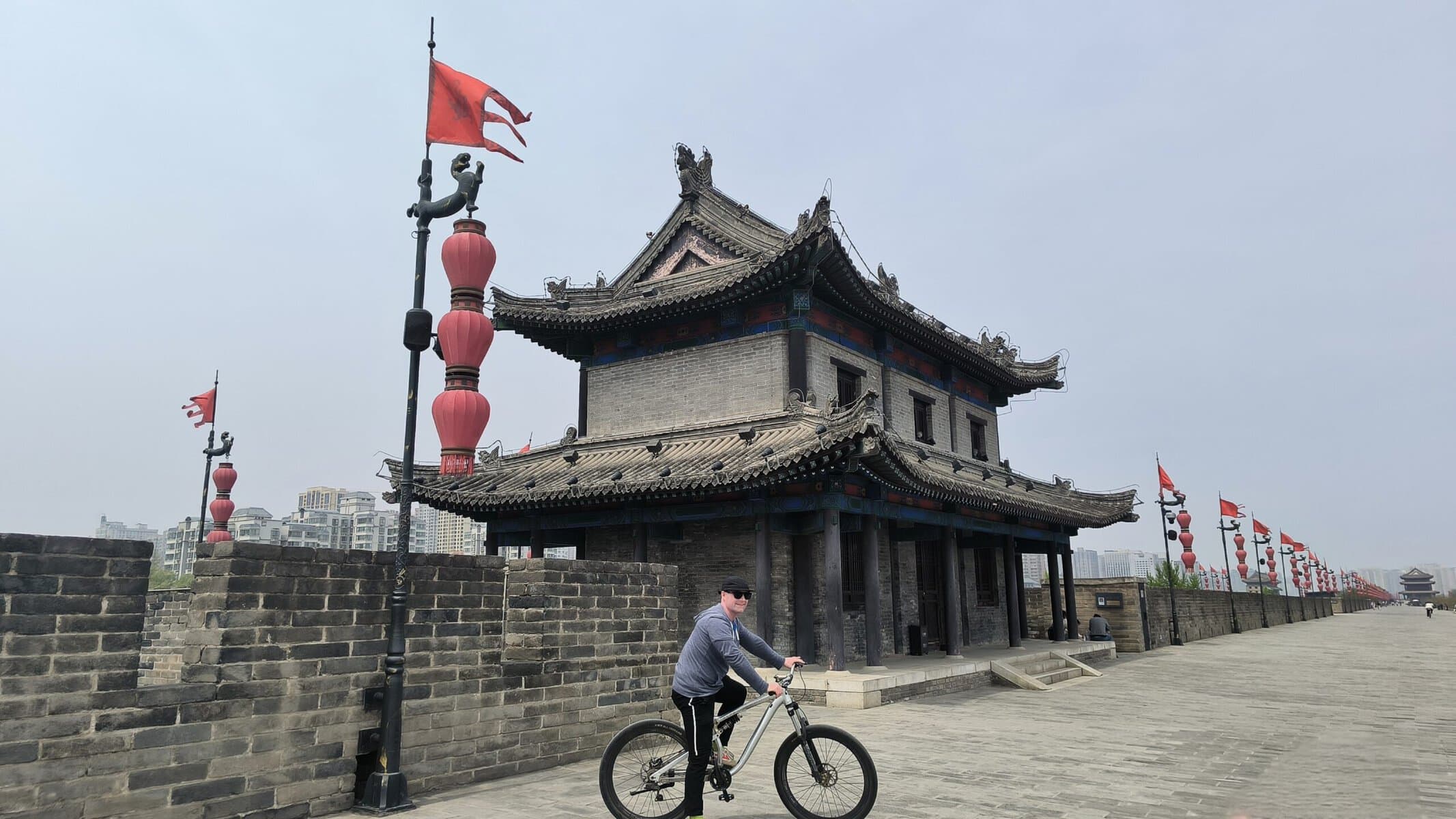 Tourists cycling atop Xi'an's historic City Wall, Chinatravel, Traditional Chinese temple-like building with ornate curved roof, red lanterns, and a cyclist passing by along a stone wall.