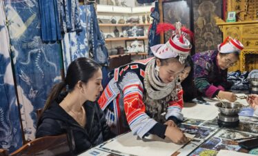 Batik experience in Miao minority, Guizhou, china, china tour, People in traditional dress at a craft workshop, writing or painting at a table with mirrors and printed sheets in a shop filled with blue fabrics and curiosities.