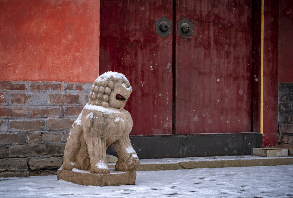 Snow-dusted stone guardian lion in front of a red wooden door with a brick base, winter scene