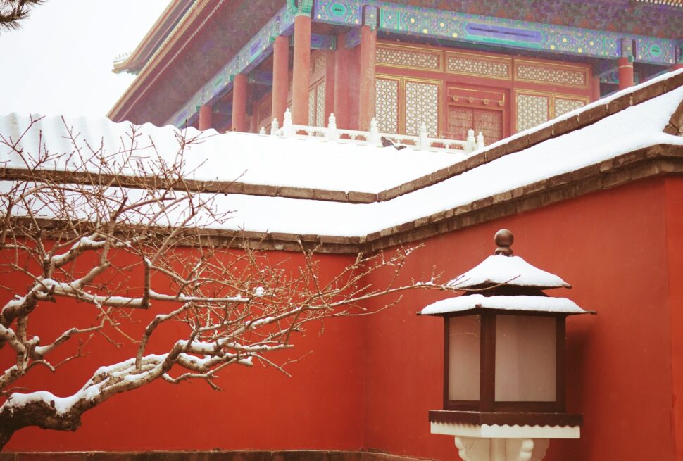Snow-covered temple courtyard with red walls and ornate multi-tiered roof; foreground white lantern and snow‑capped branches in the foreground.