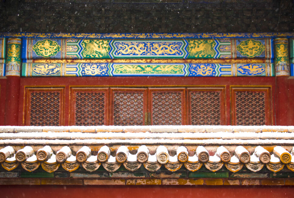 Colorfully painted temple eaves with dragon motifs in blue, green, and gold above red lattice doors; light snow on the roof tiles.