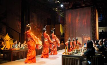 sichuan, china, china tour, Dancers in bright orange, ornate costumes perform a traditional dance on a stage while audience at tables records with phones.