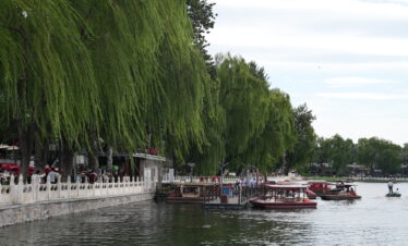 Shichahai, beijing, china, china tour, Riverside promenade with weeping willow trees, a white railing, and people walking by the water with boats docked nearby