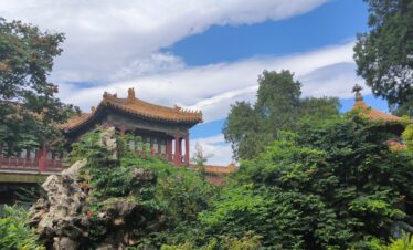 the forbidden city, beijing, china, china tour, Traditional Chinese temple with orange tiled roofs framed by lush greenery and a blue sky.Reflective garden rocks and red wooden beams visible in the foreground, surrounding the temple.