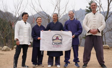 wudang taichi, hubei, china, chinaexplorertour, Five people in martial arts outfits stand outdoors holding a banner that says 'China Explorer Tour' with a mountainous, leafless-tree backdrop.