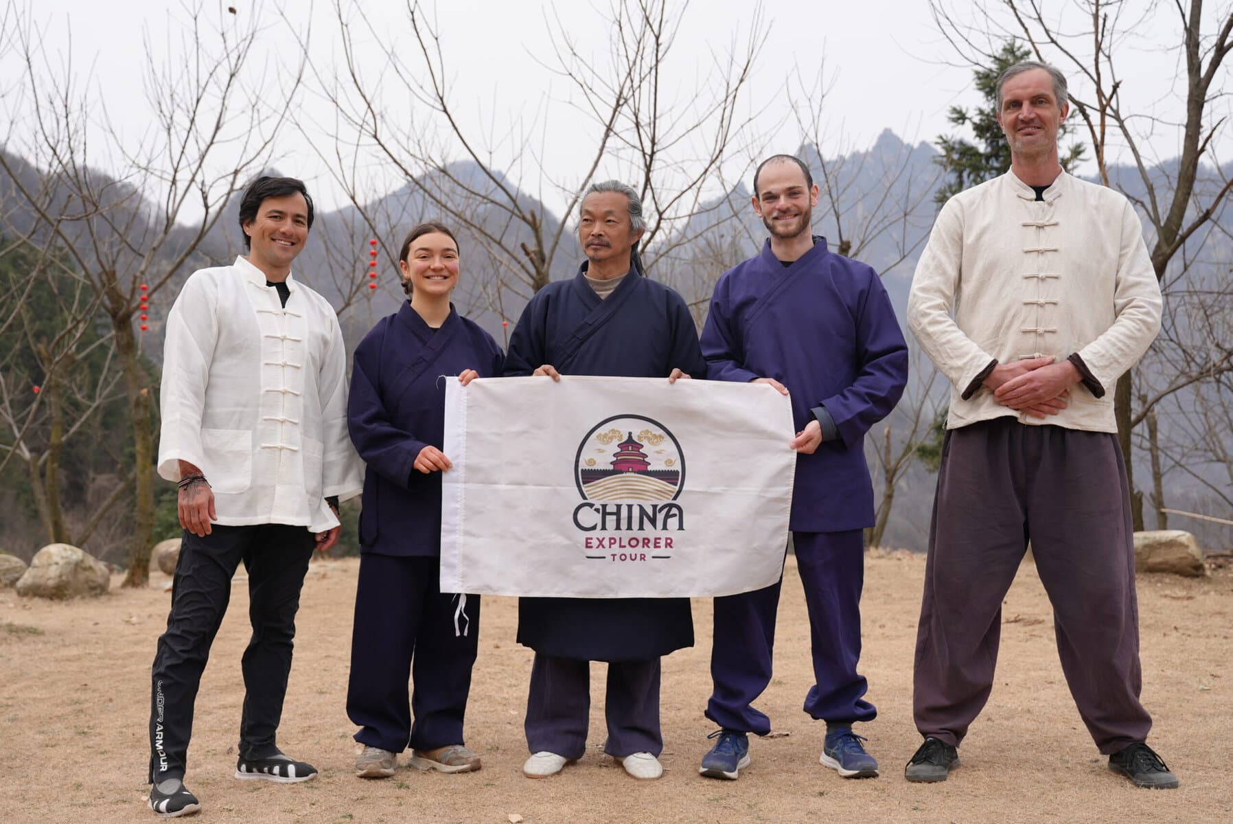 wudang taichi, hubei, china, chinaexplorertour, Five people in martial arts outfits stand outdoors holding a banner that says 'China Explorer Tour' with a mountainous, leafless-tree backdrop.