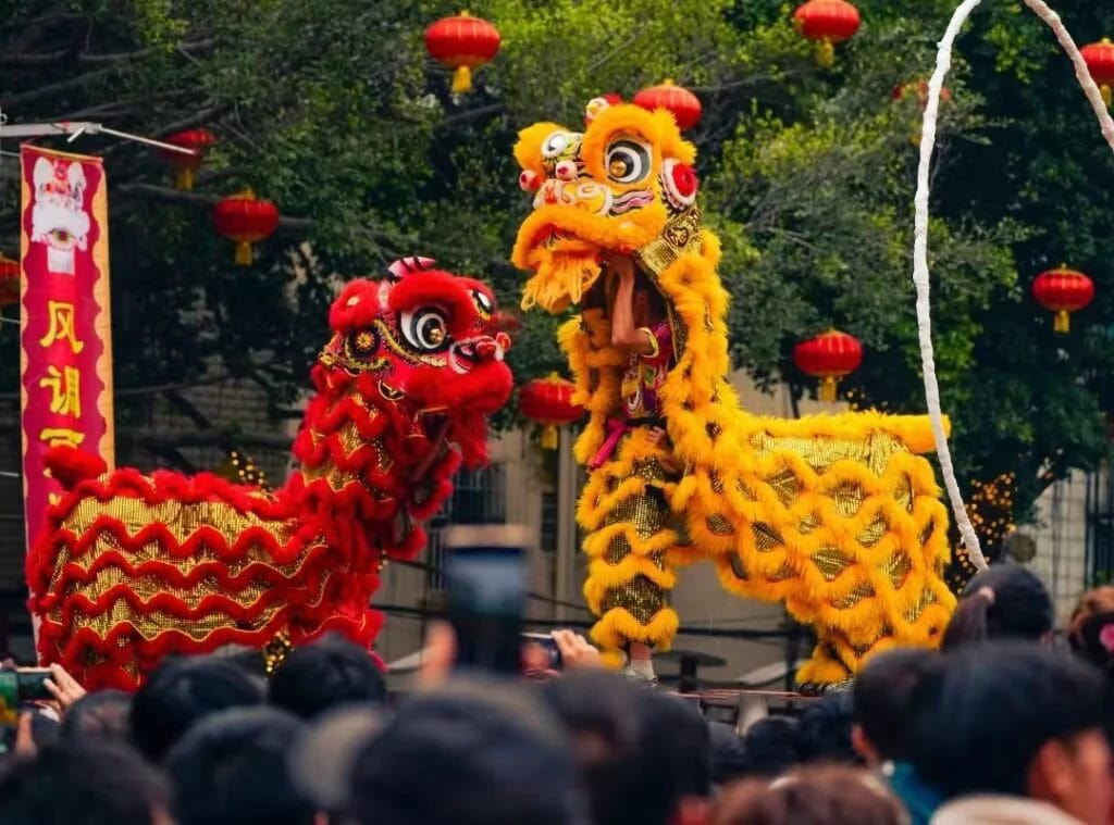 Celebrate the Chinese New Year with China Explorer Tour, Crowd watches a traditional Chinese lion dance with red and yellow lions amid red lanterns at a festive street event.