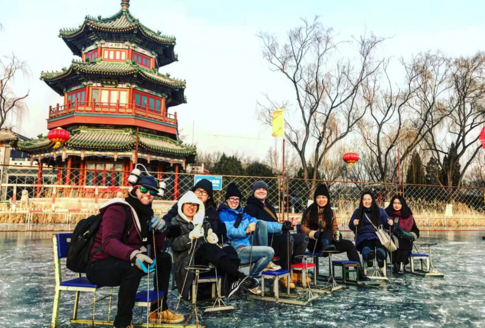shichahai ice skating rink, china, china tour, Group of friends in winter gear sitting on ice chairs on a frozen pond, with a colorful multi-tier Chinese pagoda and bare trees in the background.