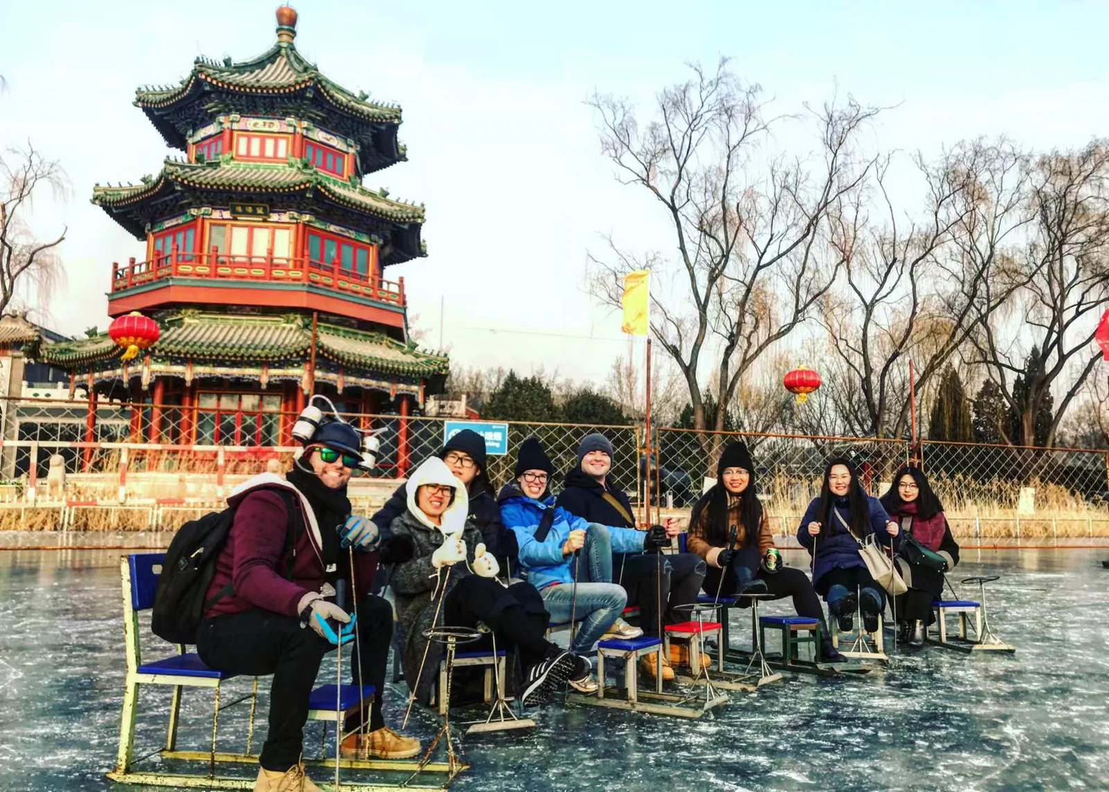 shichahai ice skating rink, china, china tour, Group of friends in winter gear sitting on ice chairs on a frozen pond, with a colorful multi-tier Chinese pagoda and bare trees in the background.