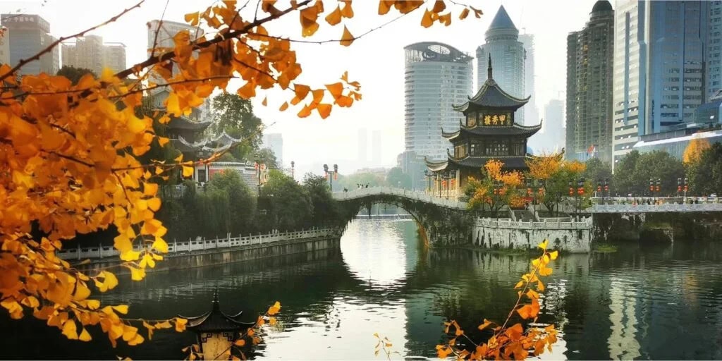 jiaxiu tower, guiyang, guizhou, china, china tour, Autumn leaves frame a calm river with a traditional Chinese pavilion and arched stone bridge, city skyscrapers in the background.