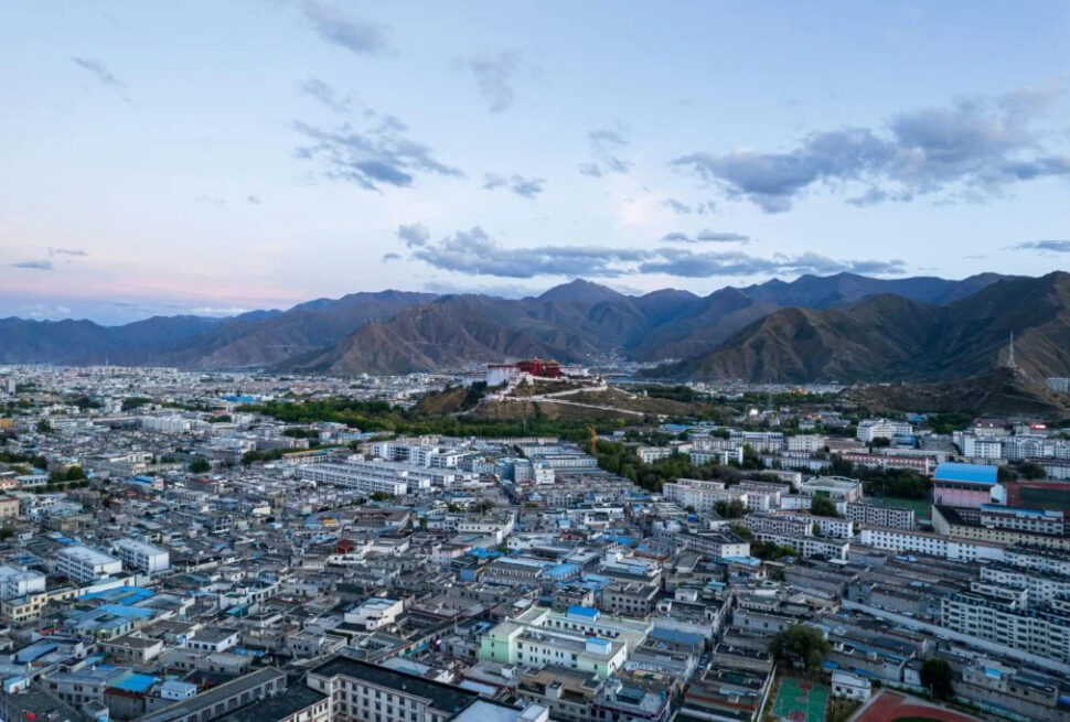 tibet, china, china tour, Aerial view of a densely built city with blue-roofed buildings and a distant mountain range under a cloudy sky.