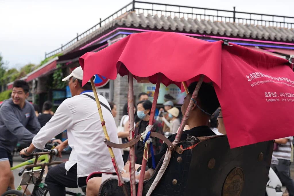 The Importance of Hutongs: Exploring Old Beijing Neighborhoods, china tour, Pedicab with a bright pink canopy carrying a rider, on a busy street with pedestrians and cyclists nearby.