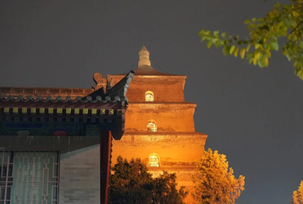 great wild goose pagoda, xi'an, china tour, Night view of a three-story brick pagoda lit from below, with a traditional roofed building in the foreground and trees on the right.