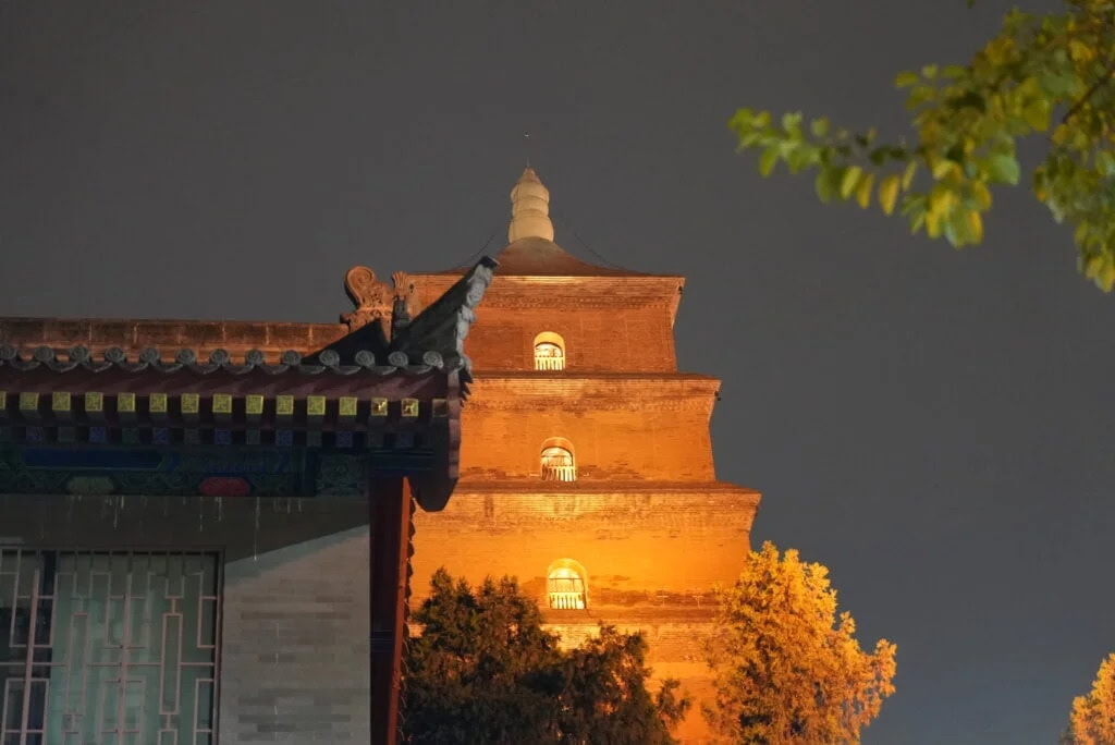 great wild goose pagoda, xi'an, china tour, Night view of a three-story brick pagoda lit from below, with a traditional roofed building in the foreground and trees on the right.