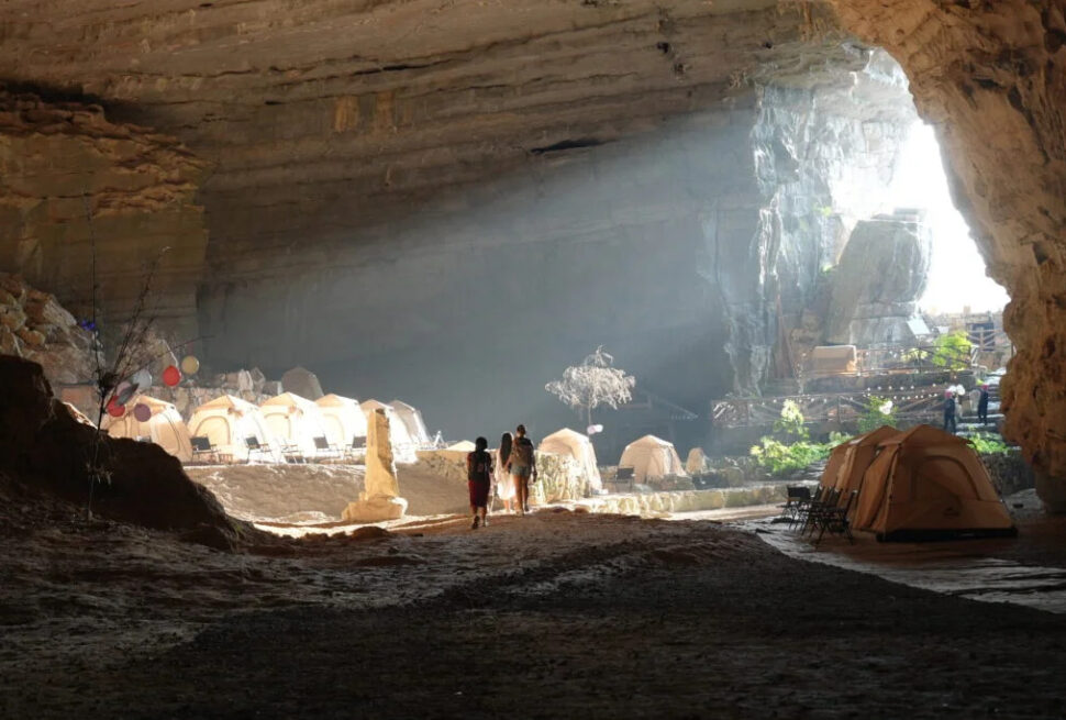guizhou cave, china, china tour, Campsite inside a large cave with beige tents, a few people walking, and sunlight at the cave entrance.