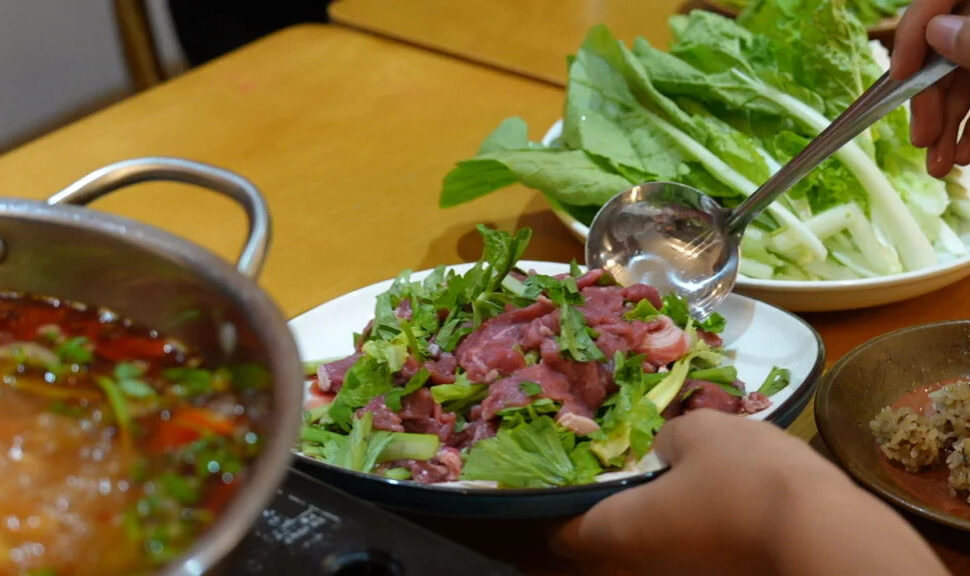 guizhou food, china, china tour, Plate of beef salad with fresh herbs being served, with leafy greens and a pot of broth nearby on a wooden table.