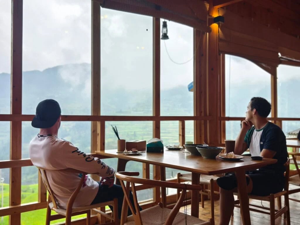 guizhou, china, china tour, Two men sit at a wooden table in a bright cafe, overlooking misty mountains through large windows.