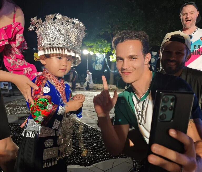 guizhou, china, china tour, Child in a silver, ornate headdress and colorful traditional costume posing for a selfie with a smiling man holding a smartphone at night.