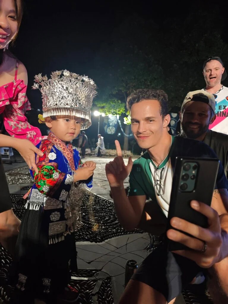 guizhou, china, china tour, Child in a silver, ornate headdress and colorful traditional costume posing for a selfie with a smiling man holding a smartphone at night.