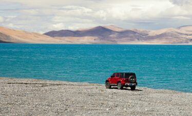 Tibet, china, china tour, Red Jeep parked on a rocky shore beside a bright blue lake with arid mountains in the distance under a cloudy sky