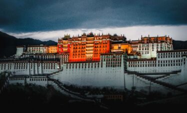 Potala Palace Tibet, china, china tour, Potala Palace perched on terraced hills with warm orange buildings under a dramatic, dark storm sky at dusk.