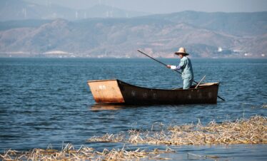 8-Day Yunnan Culinary Cultural Adventure, china, china tour, Person rowing a small rust-colored boat on a calm blue lake, wearing a wide-brimmed straw hat and light clothing, with distant mountains in the background