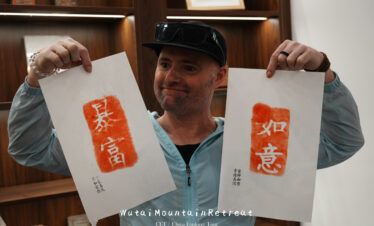 wutai retreat, shanxi, chinatrip, Smiling man in a light blue jacket holds up two calligraphy prints with orange-red seals and white Chinese characters.