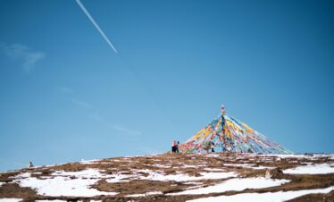 Tibet, china, china tour, Blue sky with a contrail and a hill decorated with a pyramid of colorful prayer flags on a snow-dusted slope, with a few people nearby.