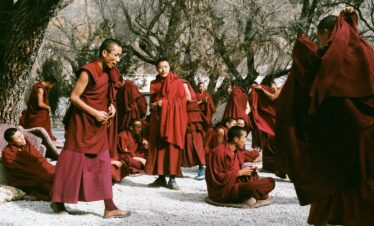 Tibet, china, china tour, Buddhist monks in maroon robes gathering outdoors among trees, some holding prayer beads on a gravel ground.
