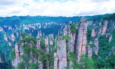 Zhangjiajie National Forest Park, hunan, china, chinatrip, Tall sandstone pillars rise from a forested canyon, lush green trees clinging to the cliffs under a cloudy blue sky.