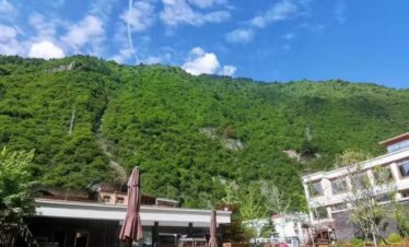 Hot Spring, Bipenggou, 3-Days Explore Western Sichuan in Winter- Siguniang Mountain, Bipenggou, and Dagu Glacier, china tour, Lakeside resort with a wooden deck, brown umbrellas, and lounge chairs by calm water, set against green mountains under a blue sky with scattered clouds