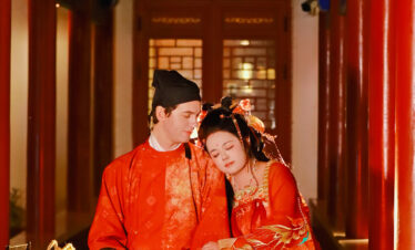 hanfu photoshoot, xingqing palace, xi'an, tang dynasty, couple, chinatravel, Couple in red traditional Chinese wedding attire sitting together in a decorated corridor, heads resting on each other under colorful beams in a temple setting.