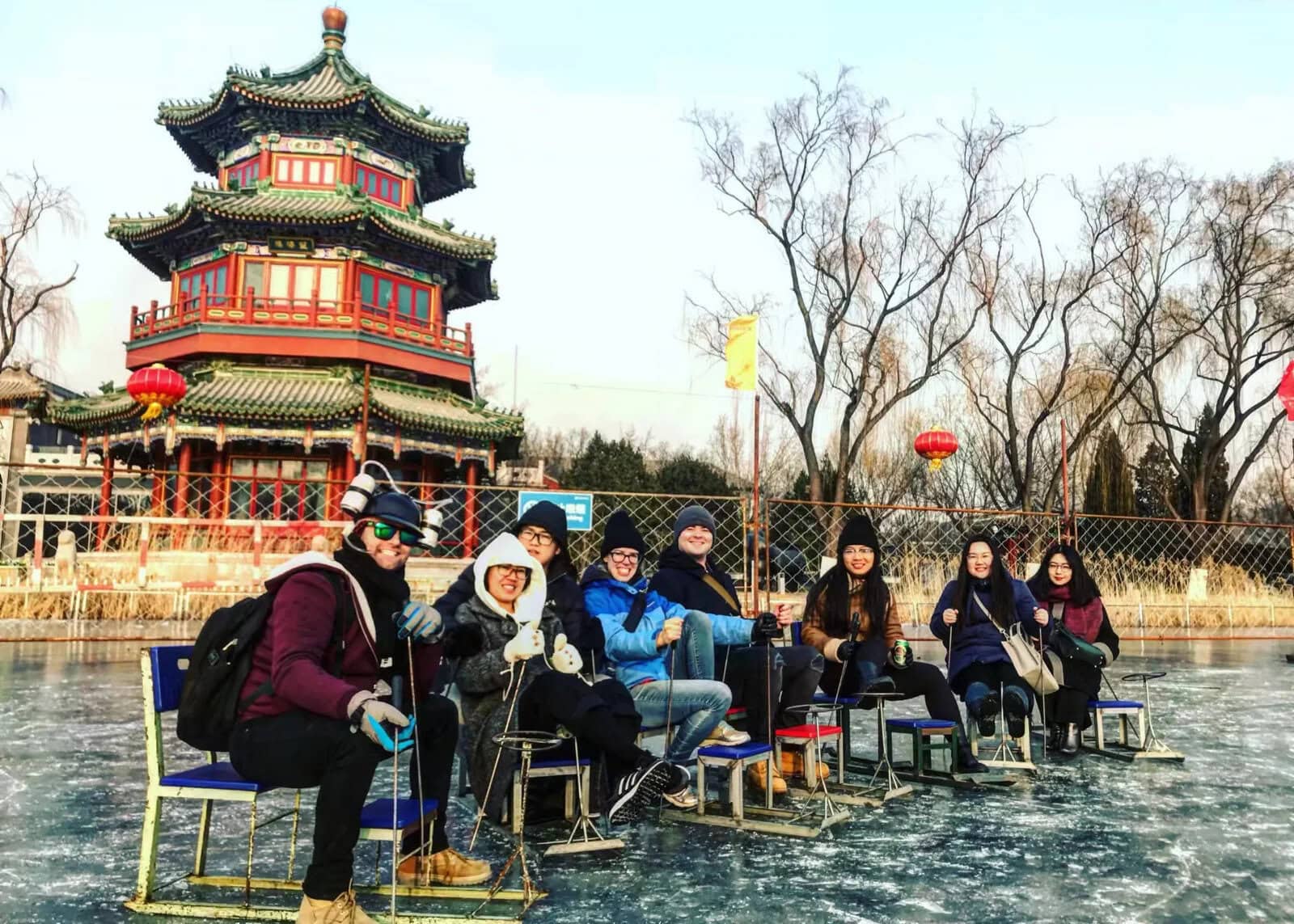 beijing pagoda scenic area, chinaexplorertour, Group of nine people in winter gear posing and smiling while seated on chairs on a frozen pond, with a traditional Chinese pagoda and red lanterns in the background.