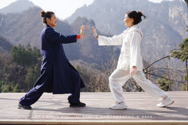 Wudang Tai Chi Retreat, hubei, china, chinaexplorertour, Two people in navy and white martial arts outfits on a wooden platform, mid high-five with a mountain landscape behind them.