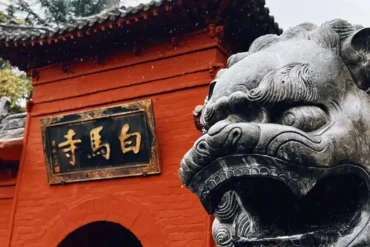 white horse temple, luoyang, henan, china tour, Red temple entrance with a carved stone guardian lion in the foreground and a gold-inscribed plaque on the wall.