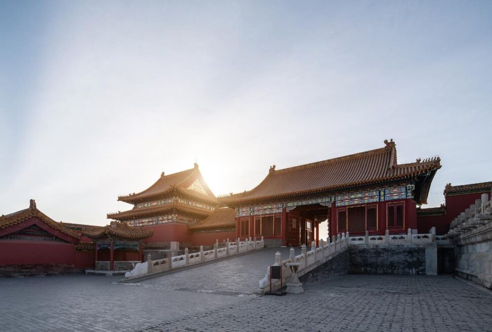 the forbidden city, beijing, china, chinaexplorertour, Ancient Chinese palace complex with red walls, ornate tiled roofs, and a stone-paved courtyard at dawn/sun behind the buildings.