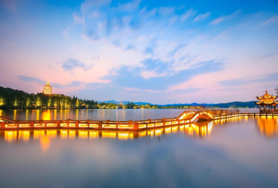 West Lake, hangzhou, zhejiang, china tour, Long-exposure view of a lit wooden boardwalk across a calm lake at twilight, with a pagoda on the left and a pavilion on the right.