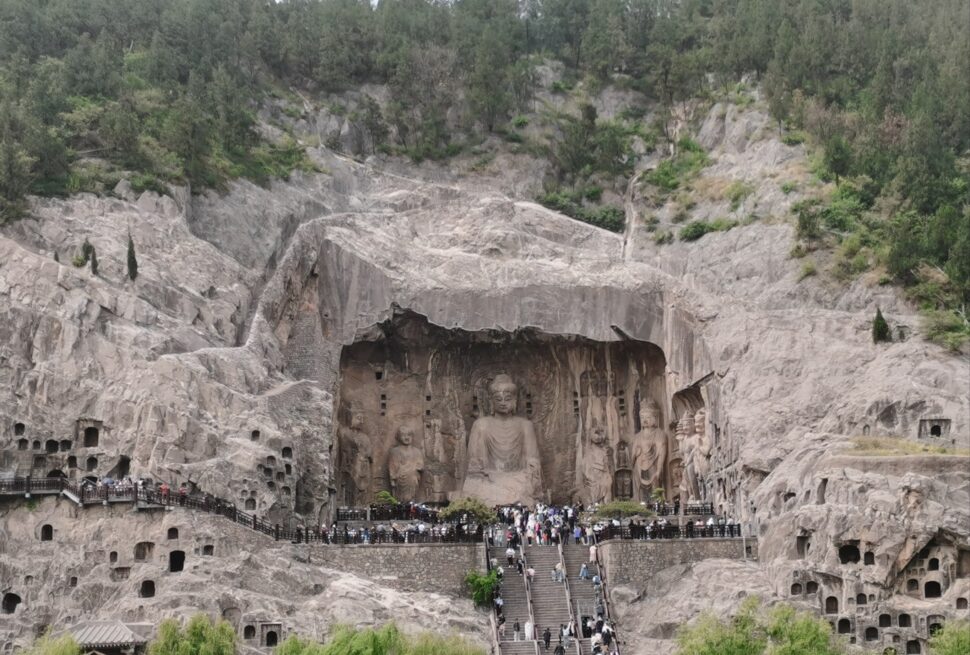 Longmen Grottoes, luoyang, henan, china tour, Rock-cut Buddhist grotto wall featuring a large central seated Buddha flanked by smaller statues, with a stone platform and railing in front., Large rock-cut Buddha statues carved into a cliff face, with a stepped walkway and crowd of visitors at the base.