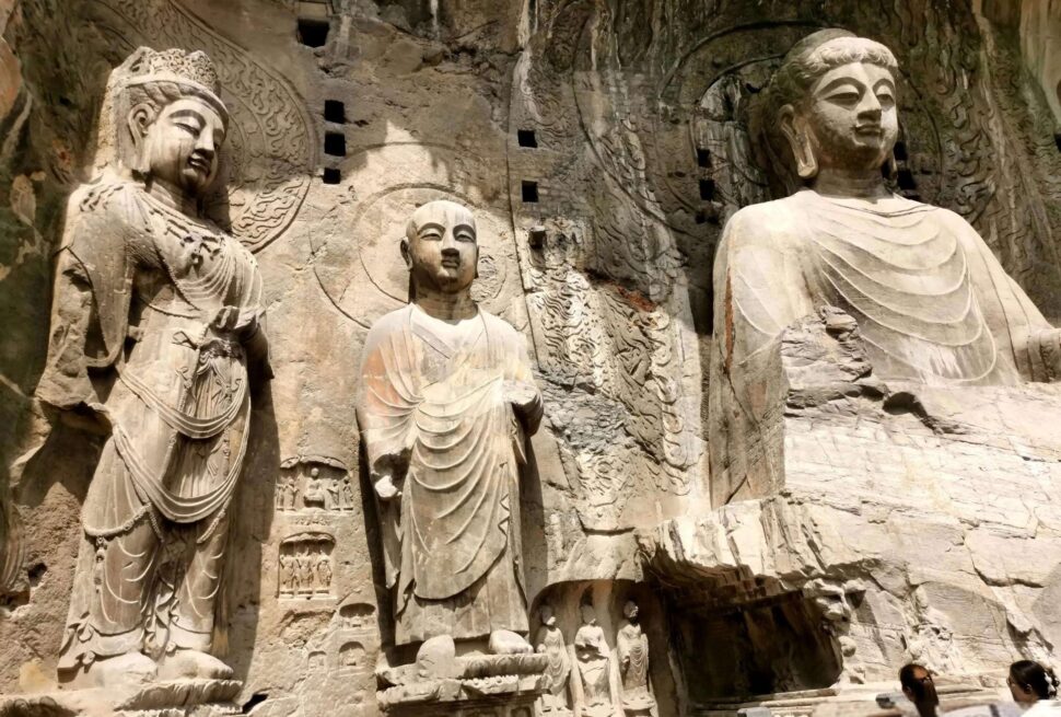 Longmen Grottoes, luoyang, henan, china tour, Rock-cut Buddhist grotto wall featuring a large central seated Buddha flanked by smaller statues, with a stone platform and railing in front., Three large stone Buddha statues carved into a rock cliff, with smaller figures at the base and intricate surrounding reliefs.
