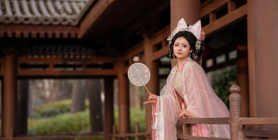 hanfu photoshoot, chinatravel, Woman in pink traditional Chinese hanfu with elaborate headpiece stands on a wooden veranda, holding a round embroidered fan.