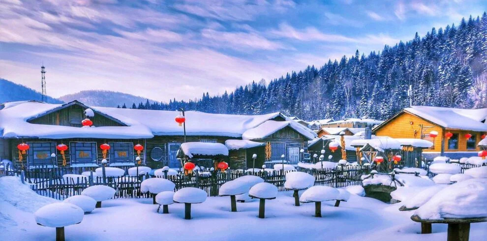 harbin, snow town, chinatravel, Snowy village scene with wooden cabins, red lanterns, and a snow-covered courtyard set against a forested hillside.