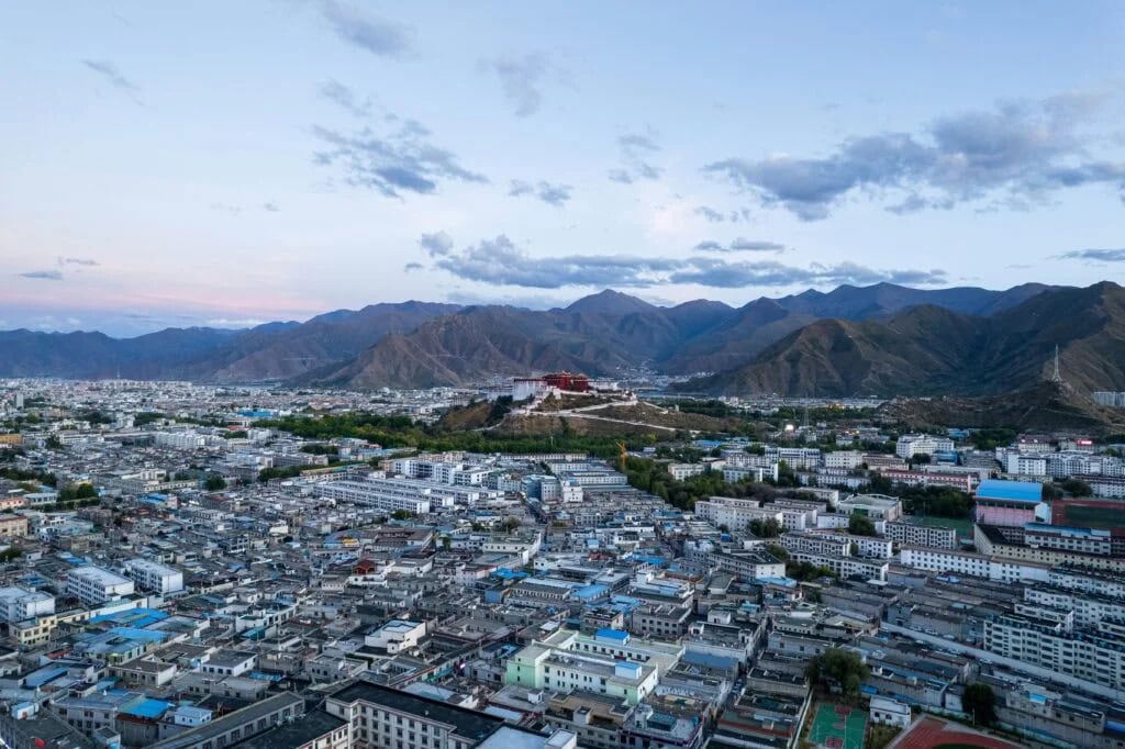 Tibet, Everest Base, Chinatravel, Panoramic aerial view of a dense city with blue-roofed buildings, set against a distant mountain range under a blue sky.