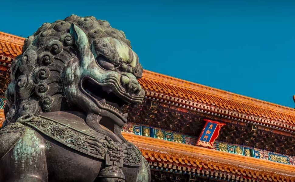 Forbidden City, beijing, china tour, Close-up of a dark stone guardian lion statue in front of a traditional Chinese temple with orange-tiled roof and colorful ornate beams against a blue sky.