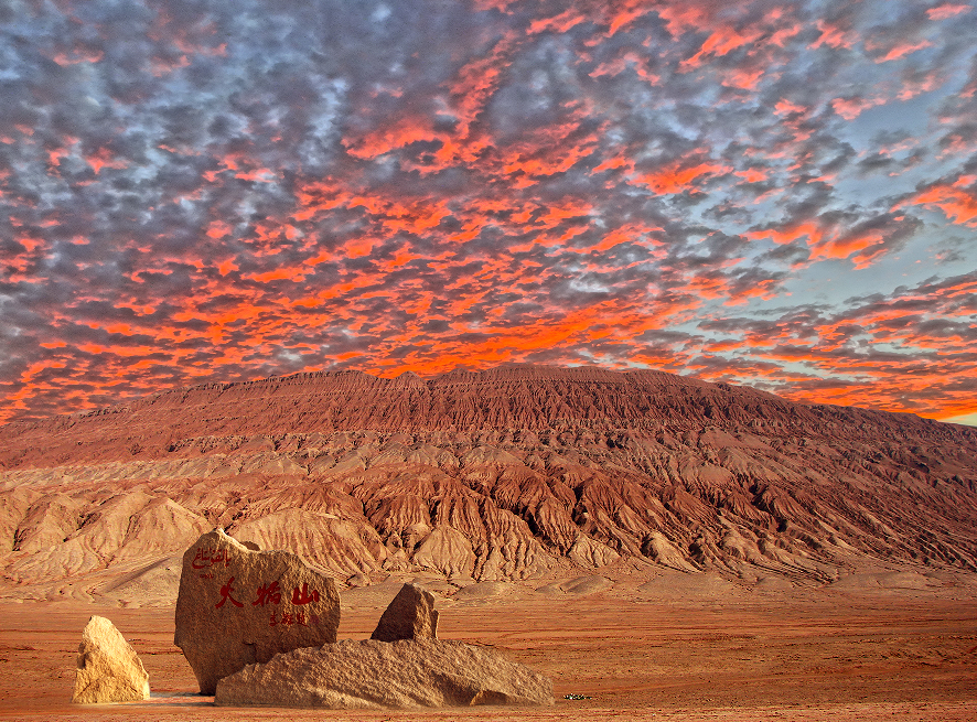 flaming mountain, turpan, xinjiang, chinatrip, Desert landscape at sunset with orange-red sky and eroded hills; a large inscribed rock and smaller boulders in the foreground.